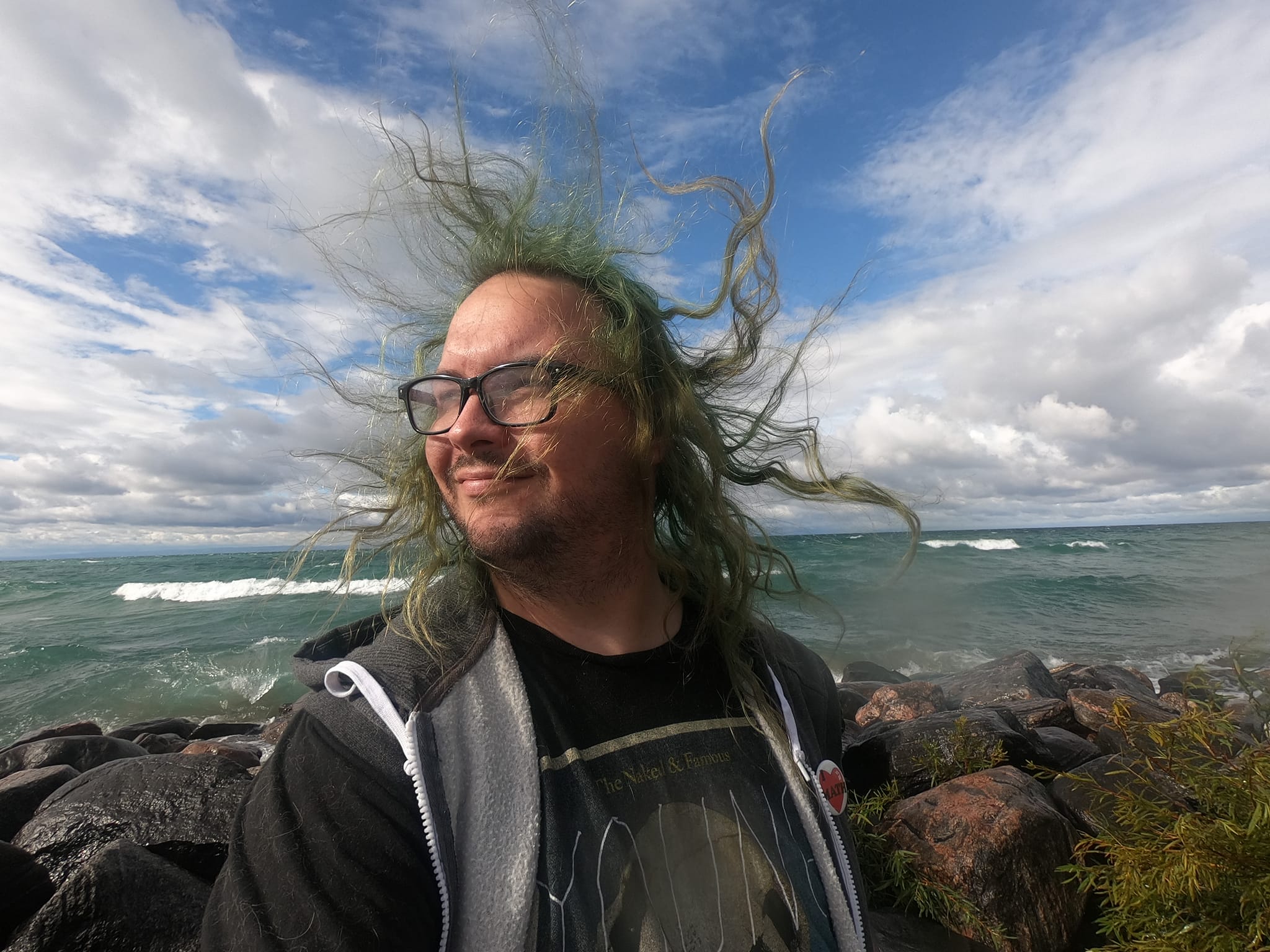 A man with long green hair wearing a hooded sweater vest stands at a breakwater, hair blowing in the wind, Photo 2